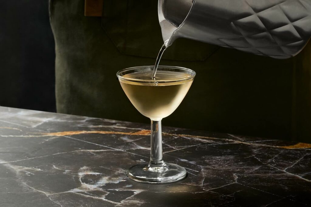 Su un piano di marmo nero con venature bianche e oro una piccola coppetta con un cocktail martini che viene versato da un mixing glass in acciaio