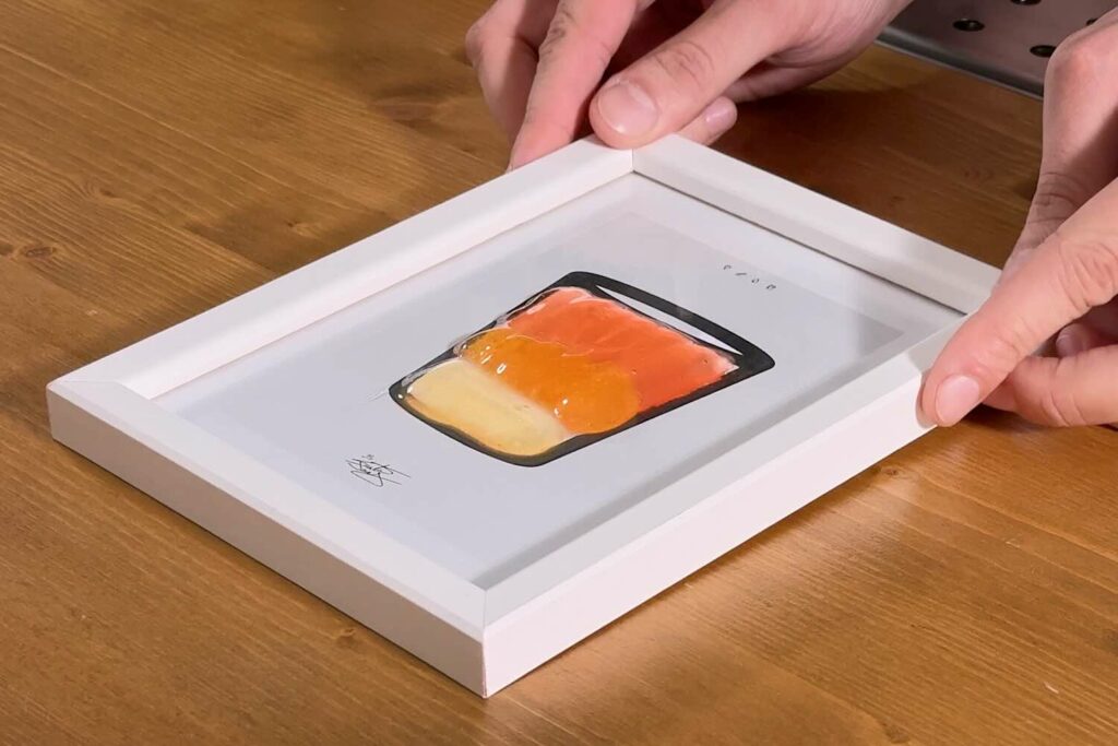 Su un banco bar in legno il "cocktail" appennello. servito su un piccolo quadro. Il vetro che protegge la stampa dell'illustrazione di un bicchiere diventa il bicchiere stesso sul quale vengono versati dei gel fluidi come pennellate.