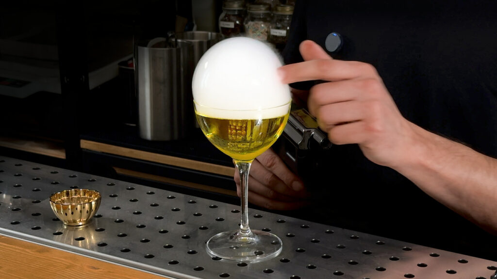 su un banco bar un cocktail giallo in una coppetta con sopra una bolla di fumo aromatico generato dalla Flavour Blaster. La bolla sta per essere scoppiata dal dito del bartender.