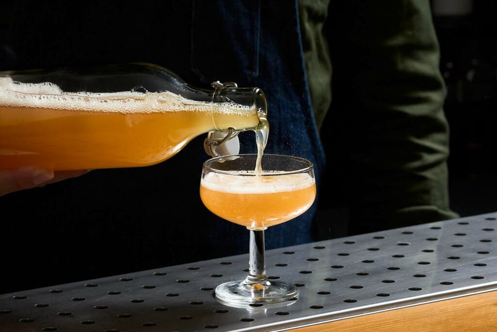 Su un banco bar, una coppetta da cocktail nella quale viene versata la bevanda fermentata "Fermentato di sciroppo di agave aromatizzata al caffè" da una bottiglia in vetro con tappo meccanico
