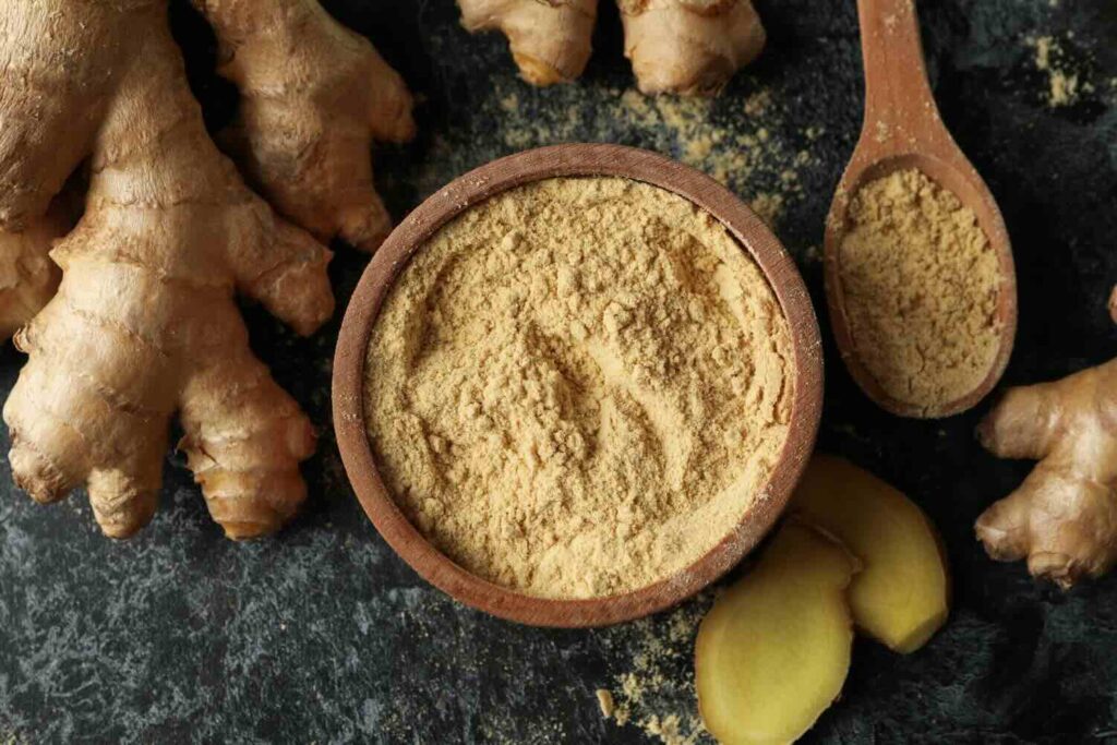 su un tavolo in marmo alcuni pezzi di zenzero fresco. Al centro una ciotola e un cucchiaio in legno con zenzero essiccato in polvere