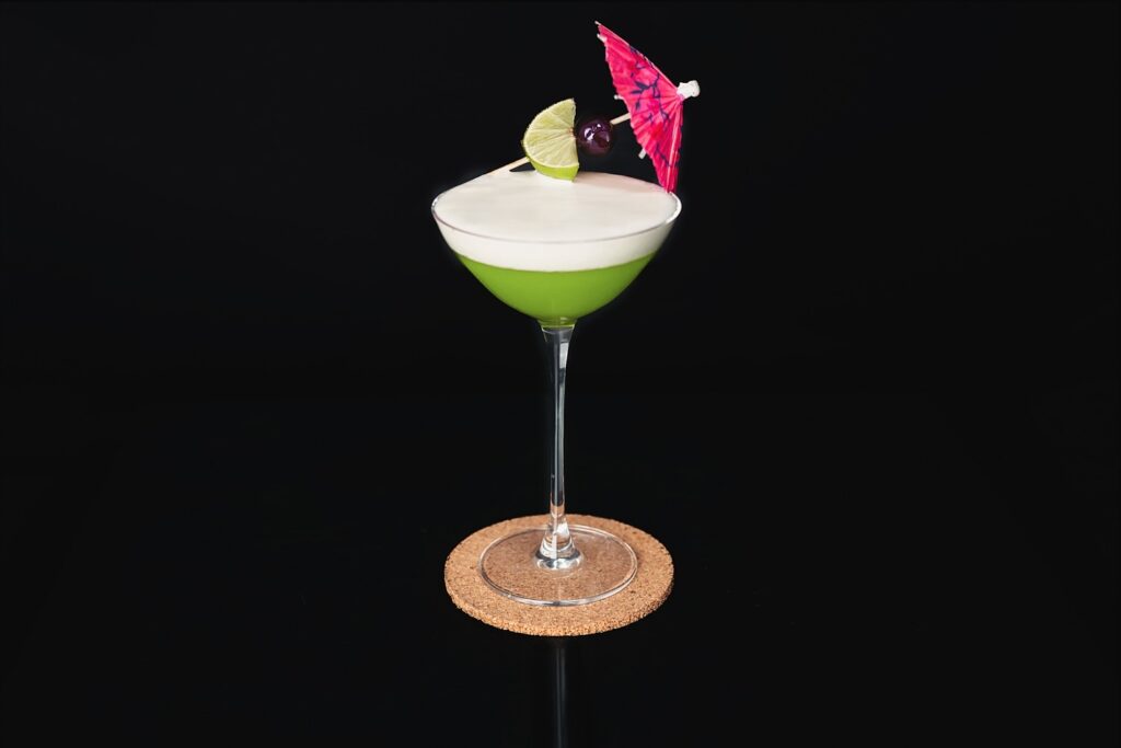 Su uno sfondo nero una coppetta da cocktail contenente il cocktail analcolico Matcha Latte Sour. Il cocktail si presenta di colore verde acceso con una foam bianca in superficie. Sulla coppetta la decorazione composta da un ombrellino, un'amarena e uno spicchio di lime