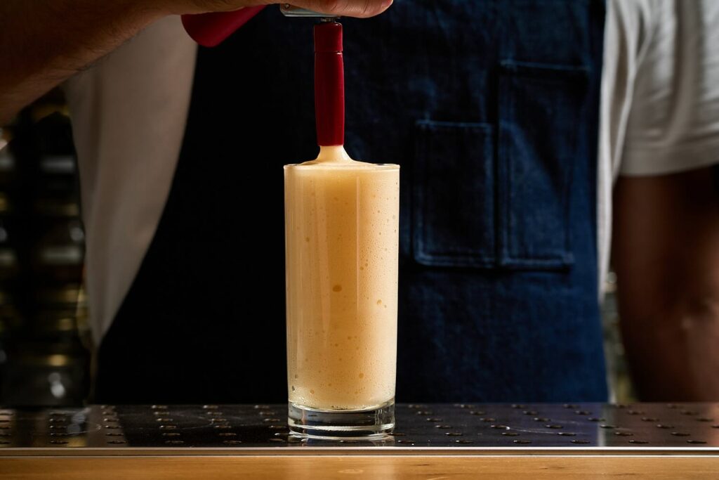 Su un banco bar un bicchiere da long drink contenente il cocktail "Bellini Molecolare" erogato da un sifone per spume