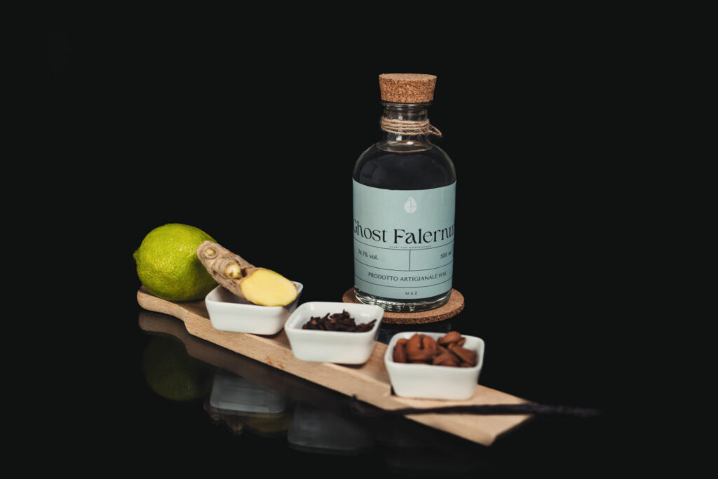 Su uno sfondo nero una bottiglia della preparazione "Ghost Falernum". davanti alla bottiglia un tagliere lungo e stretto con sopra delle ciotole bianche in ceramica contenenti zenzero, chiodi di garofano, mandorle e 1 lime
