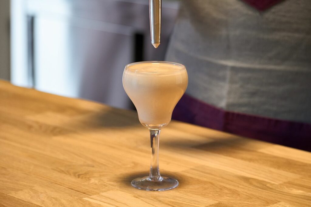 Su un banco in legno una bicchiere tipo coppetta da cocktail dalla forma allungata e sinuosa che contiene la foam calda di crêpe alla nutella. Sopra la coppetta si vede la parte terminale del beccuccio erogatore del sifone.