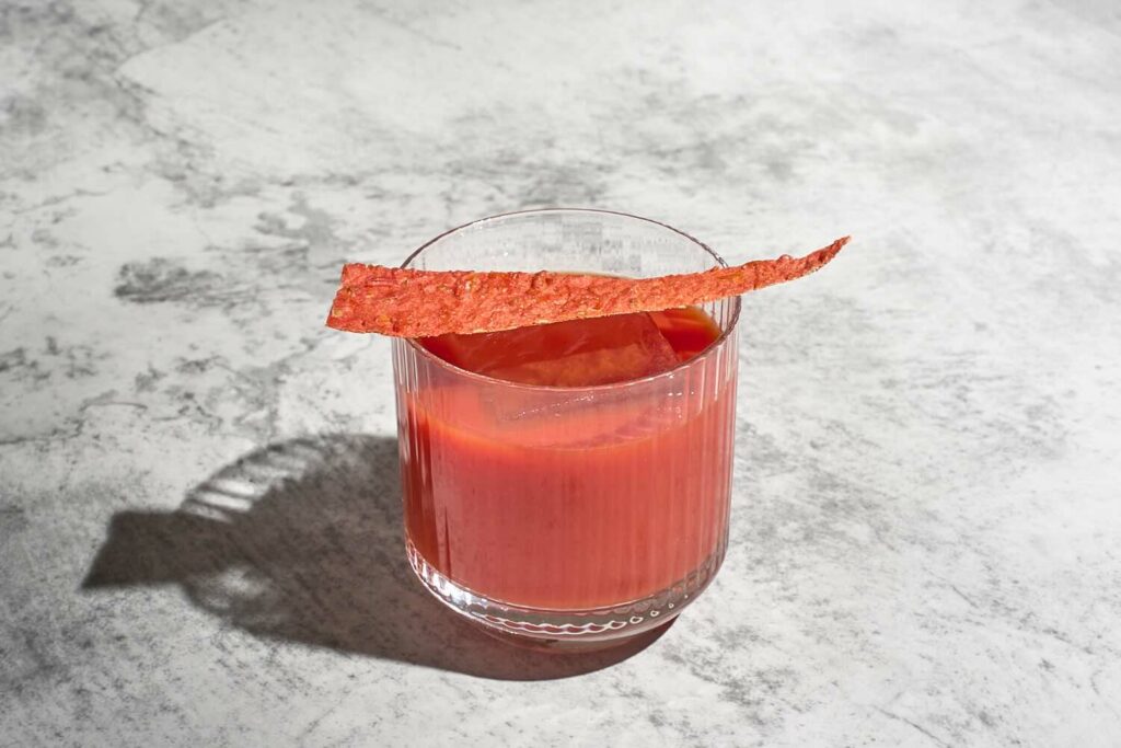 Su un piano in marmo un tumbler basso rigato con all'interno il cocktail Ho una fissa. Il drink è servito su un chunk di ghiaccio ed è decorato con un cracker di pomodoro essiccato a forma triangolare