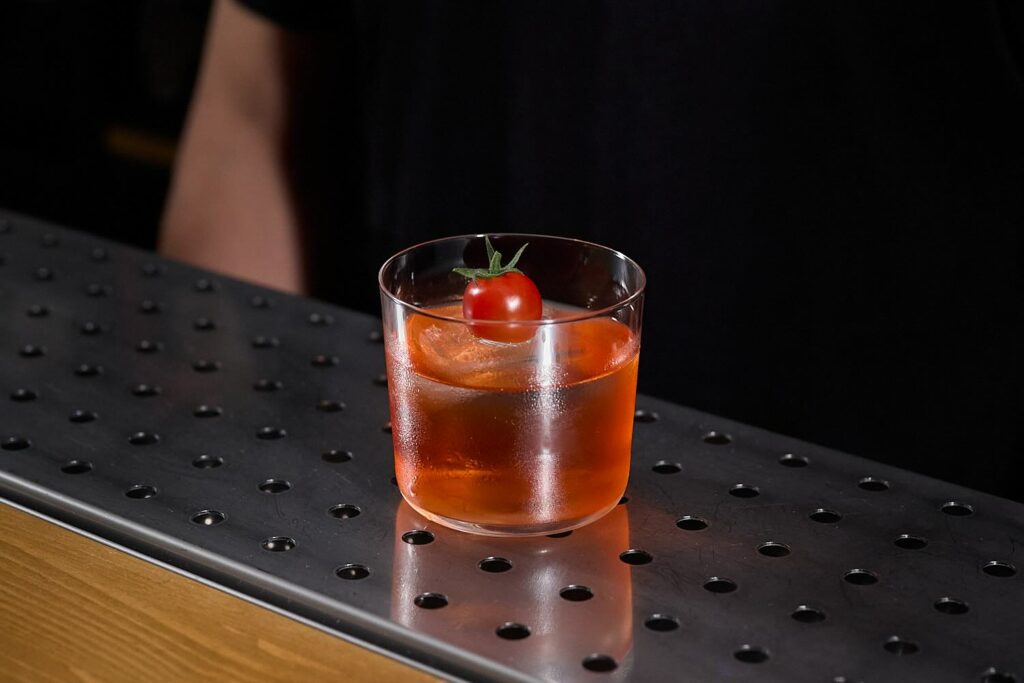 Su un banco bar un cocktail rosso servito in un tumbler basso. Il cocktail è servito su un grosso cubo di ghiaccio sul quale è posato un pomodorino rosso come decorazione.