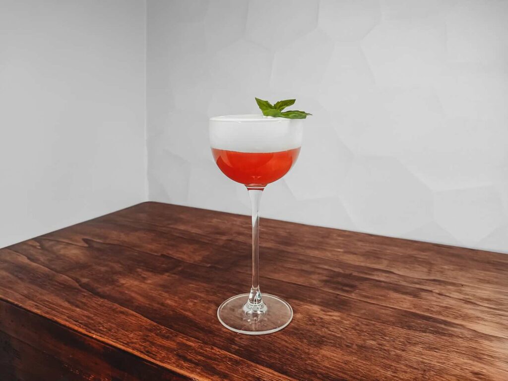 Su un tavolo di legno il cocktail bufala & pepperoni snapper. Il cocktail è servito in una coppetta da cocktail con lo stelo lungo. Si presenta di colore rosso con una foam bianca alla mozzarella di bufala. Come decorazione un ciuffetto di basilico