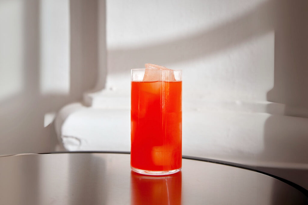 Su un tavolo con la superficie a specchio un bicchiere da long drink contenente Capsicum. Si tratta di un cocktail dal colore rosso vivo preparato con Mezcal Espadin e un premix di peperone rosso lavorato con enzimi (cellulasi e pectinasi). Il cocktail è servito su ghiaccio ed è privo di decorazione.