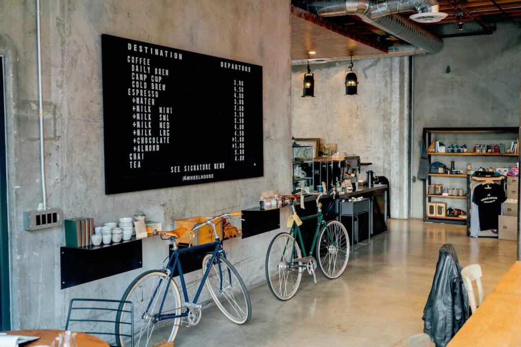 Un bar dallo stile industriale con pareti in cemento grezzo e un’illuminazione calda. Un grande menu nero con scritte bianche elenca le bevande disponibili, come caffè, tè e cioccolata calda. Due biciclette vintage, una blu e una verde, sono appoggiate contro il bancone. Sullo sfondo, una scaffalatura espone tazze, caffè in vendita e alcuni articoli di merchandising. L’atmosfera è accogliente e moderna, con un mix di elementi minimalisti e dettagli retrò