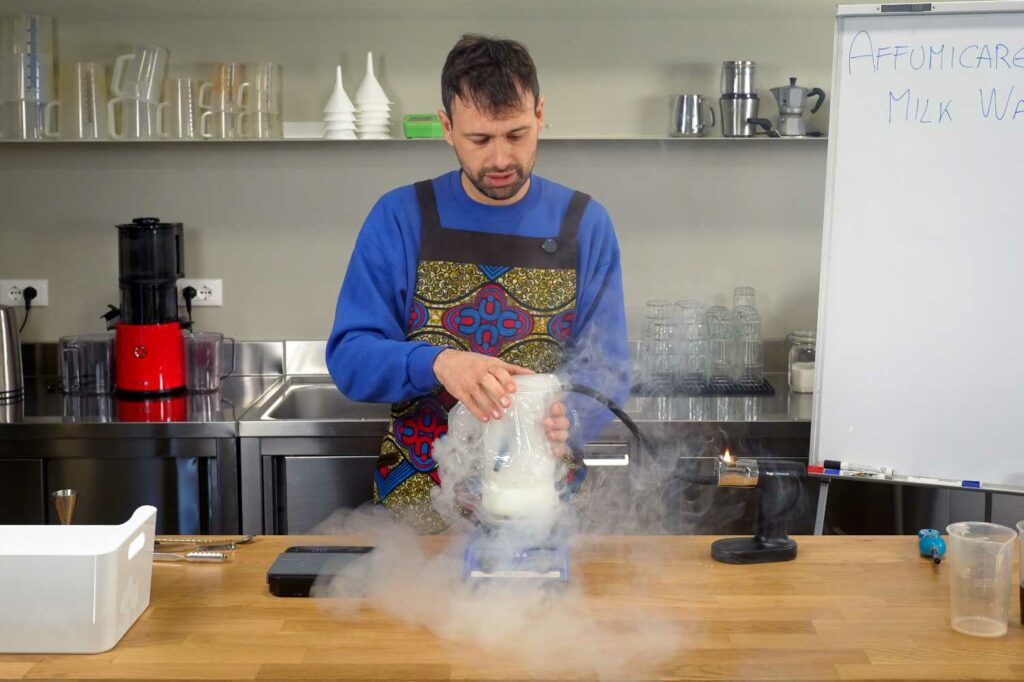 Sul bancone di un laboratorio, Giovanni Ceccarelli che sta affumicando (conuna smoking gun) del latte all'interno di una caraffa in vetro coperta con una pellicola.la caraffa è sistemata su un agitatore magnetico.