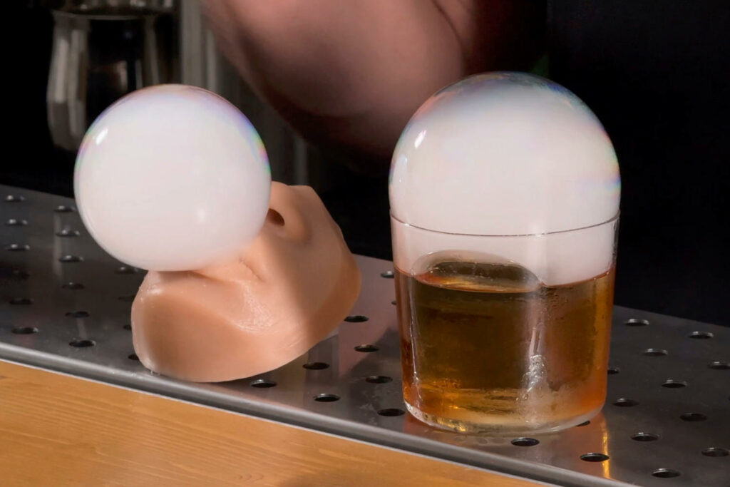 su un bancone di un bar un cocktail servito in due diversi contenitori. Uno è un volto di una ragazza in plastica alimentare e rappresenta il realismo. Il cocktail si beve dalle labbra della ragazza simulando un bacio. L'altro contenitore è un normale bicchiere tumbler basso con un chunk di ghiaccio che rappresenta astrattismo e minimalismo. I due cocktail sono decorati con una bolla di fumo aromatico erogata dalla flavour blaster.