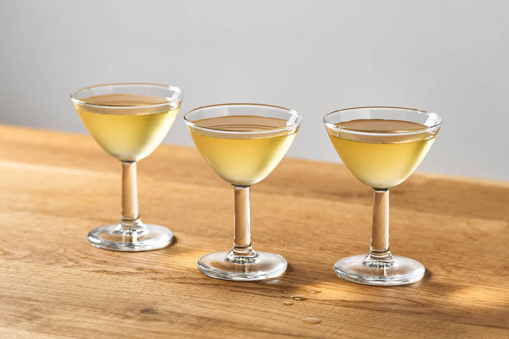 su un tavolo di legno 3 piccole coppette da cocktail contenenti 3 versioni di martini cocktail (secco, semisecco e morbido) bilanciati con Sorbole, bitter al limone.