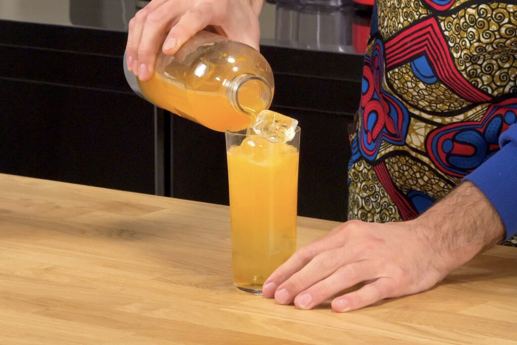 Su un tavolo in legno un cocktail long drink con Sorbole, mandarino, clairin communal e limone. Si intravede la mano del bartender che sta completando il cocktail con una soda al mandarino home-made versata da un twist'n sparkle