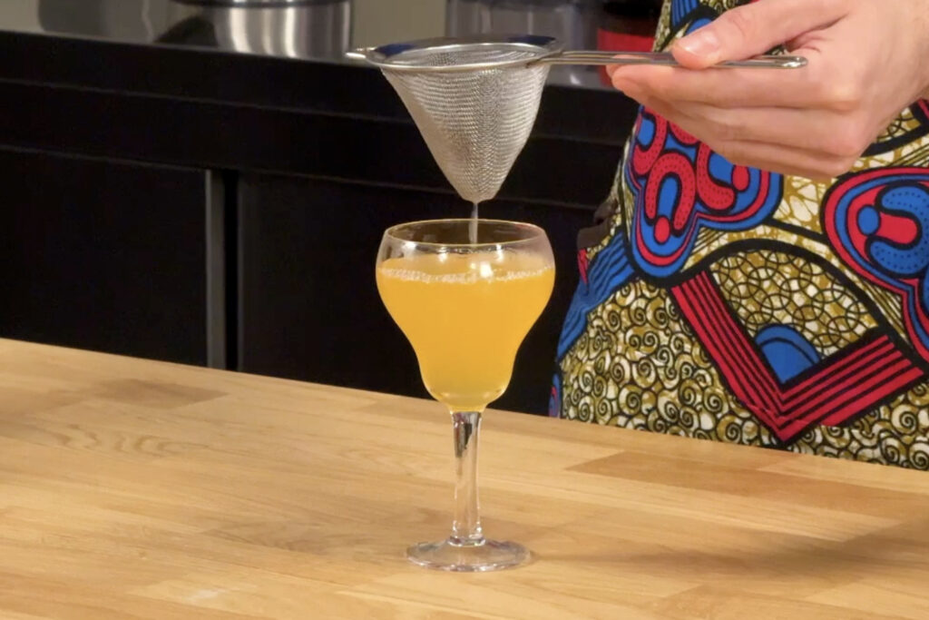 Su un tavolo in legno un cocktail con Sorbole, mandarino, clairin communal e limone. Si intravede la mano del bartender che sta filtrando il cocktail dentro la coppetta con un colino a maglie fini.