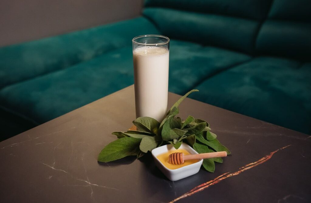 su un tavolo di marmo nero il cocktail LattePiù preparato con Rum giamaicano overproof, kefir, premix all'arancia e miele alla salvia. Sul tavolino accanto al bicchiere, un mazzetto di salvia e una ciotola con del miele