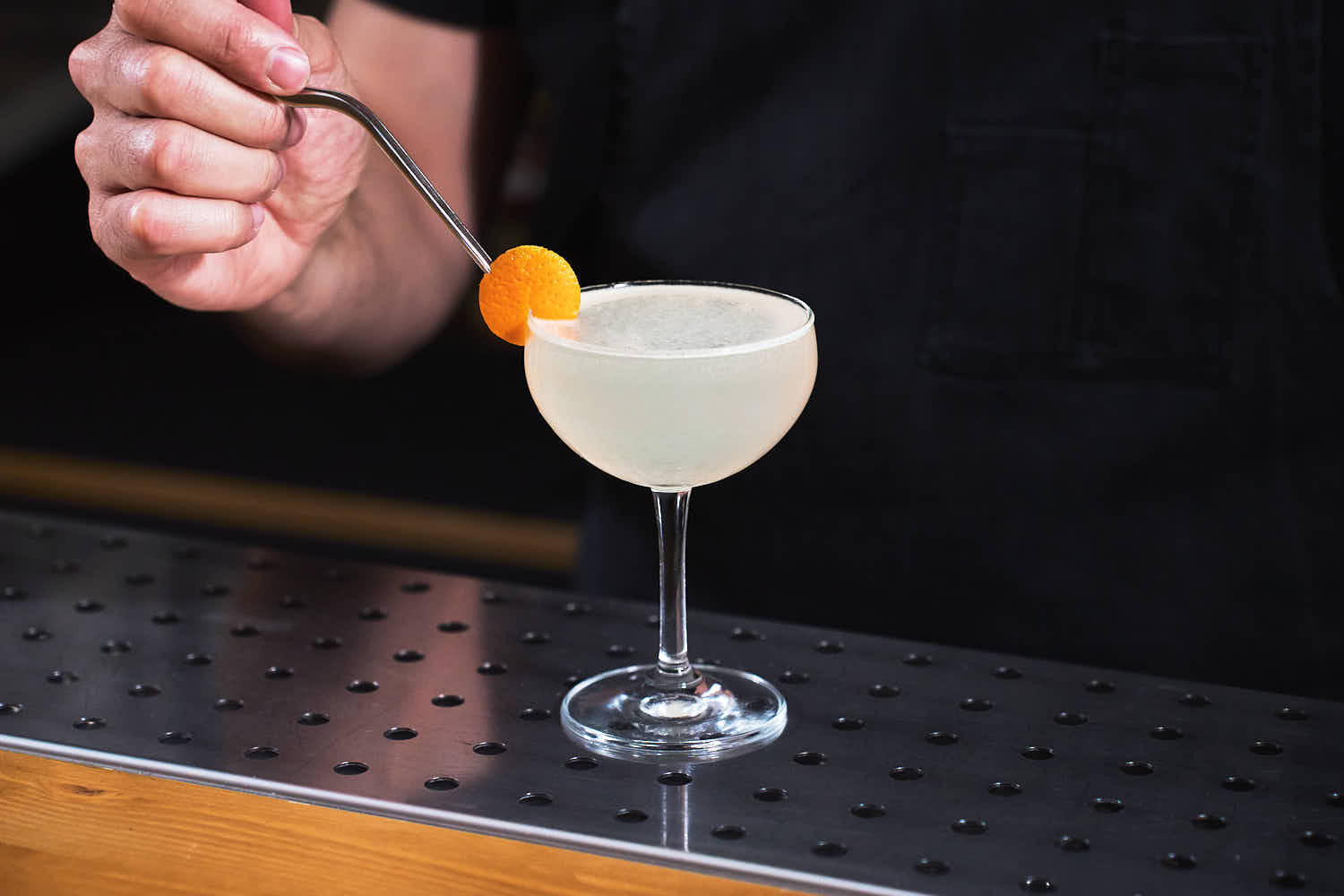 Su un banco bar una coppetta da cocktail contenente il cocktail Corpse Reviver n°2 preparato con Gin, americano cocchi, succo fresco di limone, triple sec e assenzio. Il cocktail è decorato con una scorza di arancia tagliata tonda e incastrata sul bordo del bicchiere.