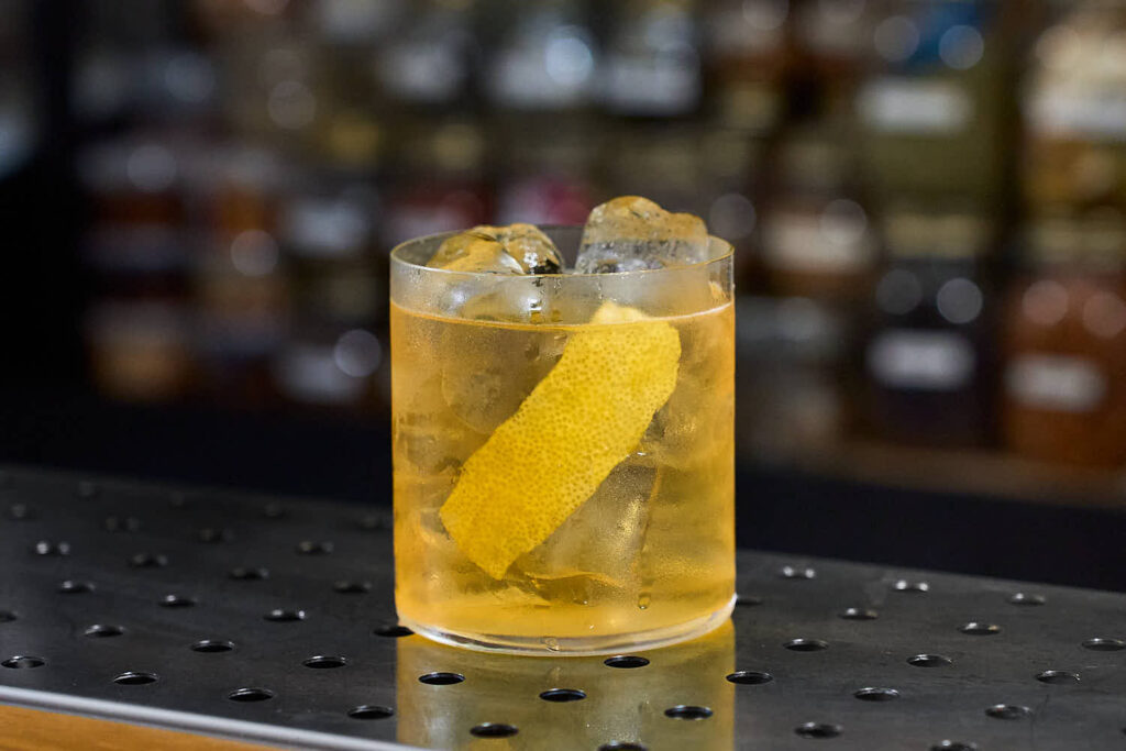 su un banco bar il cocktail "Scirocco" preparato con Bitter D'Stilla e liquore ai fiori di sambuco. Il cocktail è servito in un tumbler basso con ghiaccio e decorato con una scorza di limone