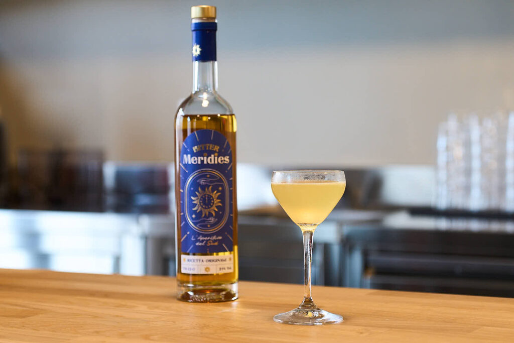 Su un banco in legno il cocktail "Terroni e Famosi", twist sul Naked & Famous preparato con Cachaça, bitter Meridies, Liquore Strega, Succo di Lime fresco e servito in una coppetta da cocktail classica. Sullo sfondo una bottiglia di bitter Meridies