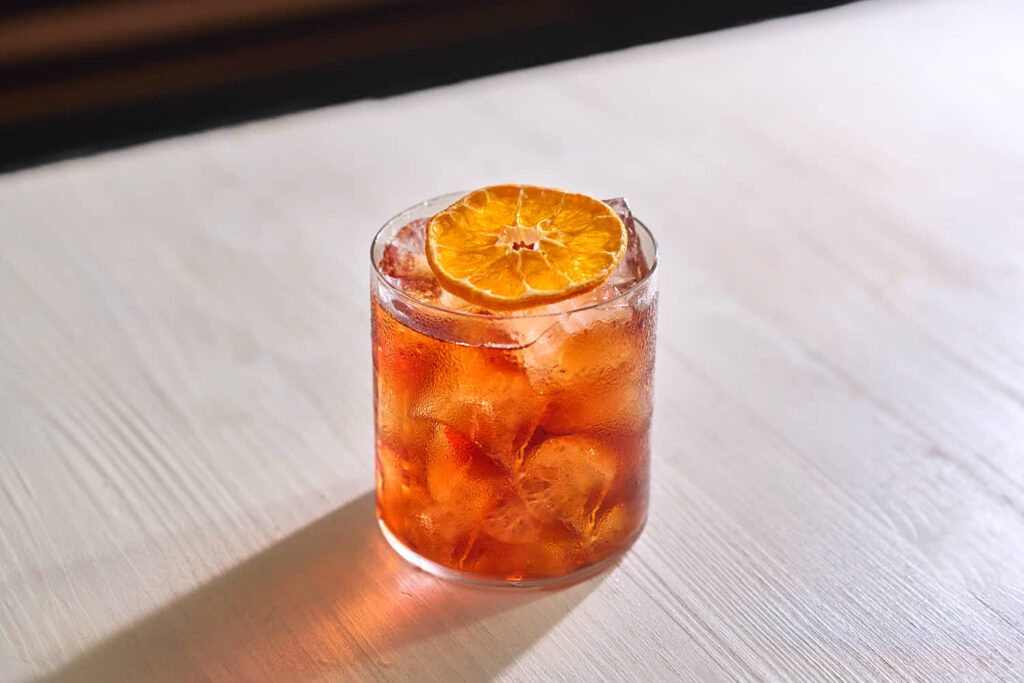 Su un piano di legno bianco il cocktail "Un Americano a Bologna" preparato con Sorbole, Bitter, Vermputh del Professore rosso e soda. Il cocktail è servito con ghiaccio in untumbler basso ed è decorato con una fetta di arancia essiccata