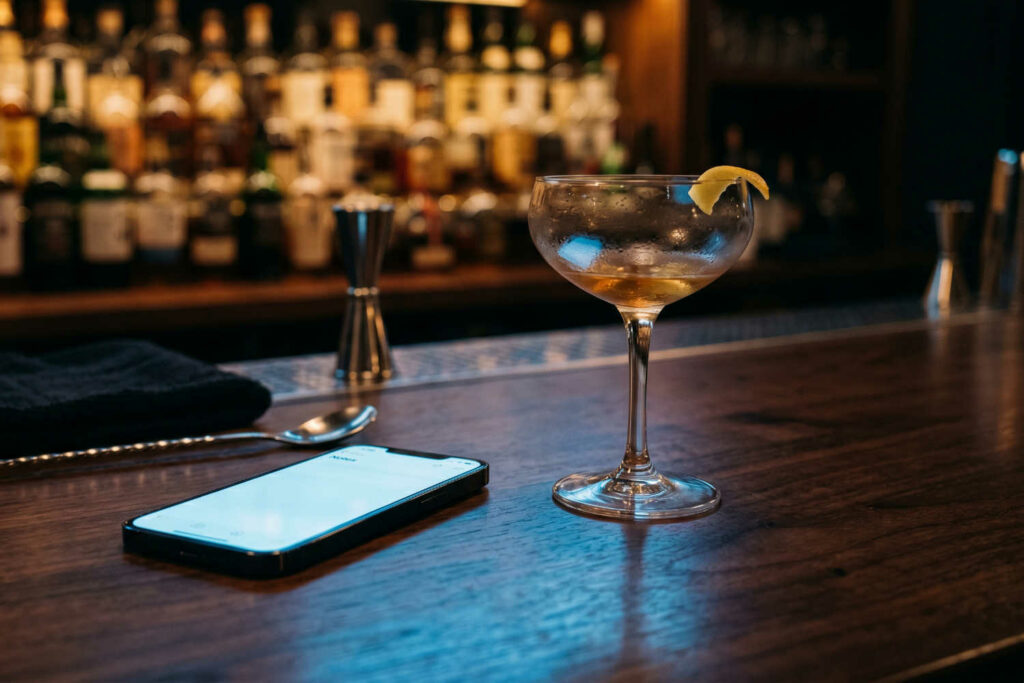 Il bancone di un bar con sopra uno smartphone e un cocktail in coppetta. Sullo sfondo sfocato si intravede la bottigliera