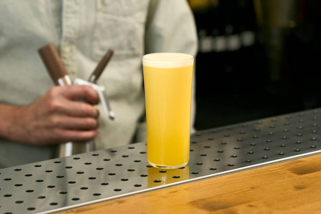 Su un banco bar il cocktail Bahia Nitro. Preparato con Succo fresco di ananas, sciroppo di acqua di cocco verde e lime. Sullo sfondo la mano del bartender che tiene il sifone iSi Nitro usato per erogare il cocktail.