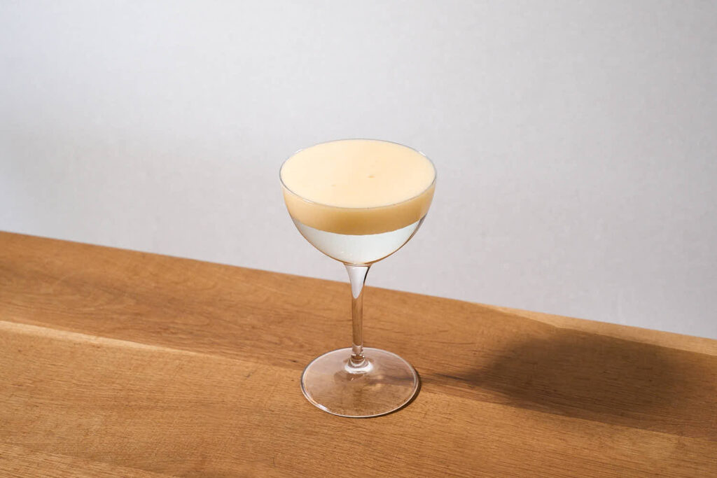 Su un tavolo di legno, una coppetta da cocktail contenente una Caipirinha rivisitata in chiave moderna. Questo cocktail è preparato con Cachaça, succo di lime chiarificato e viene servito con una foam frizzante al passion fruit in superficie.
