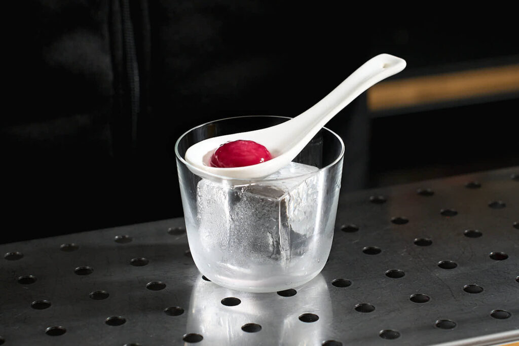 Su un banco bar la decorazione sferificata "camilla" preparata con arancia, carota viola e mandorle. La sfera è all'interno di un cucchiaio in ceramica bianco posato sopra al chunk di ghiaccio dentro al bicchiere tipo tumbler basso.