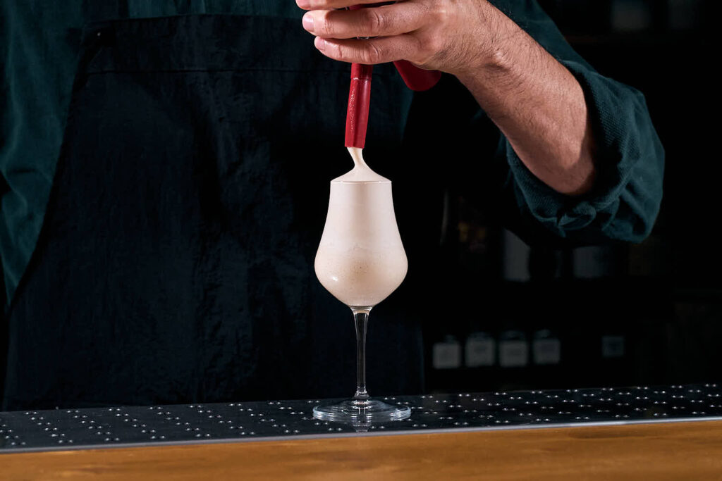 su un banco bar la mano di un bartender che tiene in mano un sifone ed eroga una schiuma gassata di coca cola all'interno di un piccolo calice svasato