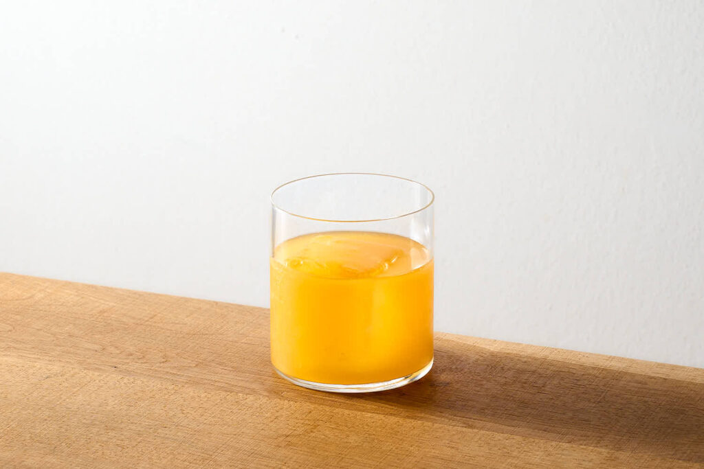 Su un tavolo di legno, in cocktail "Mango Mez" servito con un Chunk di ghiaccio, in un bicchiere tumbler basso dalla linea minimale. gli ingredienti del cocktail sono: Mezcal, acido citrico, succo di mango trattato con enzimi, succo fresco di passion fruit e zucchero