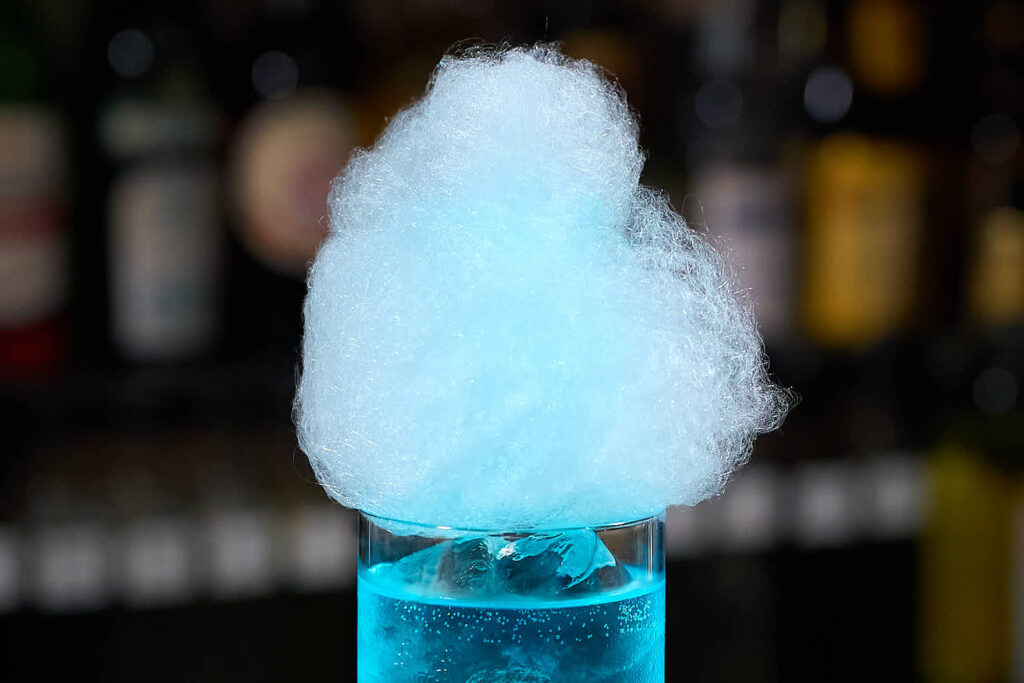 una nuvola di zucchero filato azzurro posta come decorazione su un bicchiere che contiene un cocktail blu.