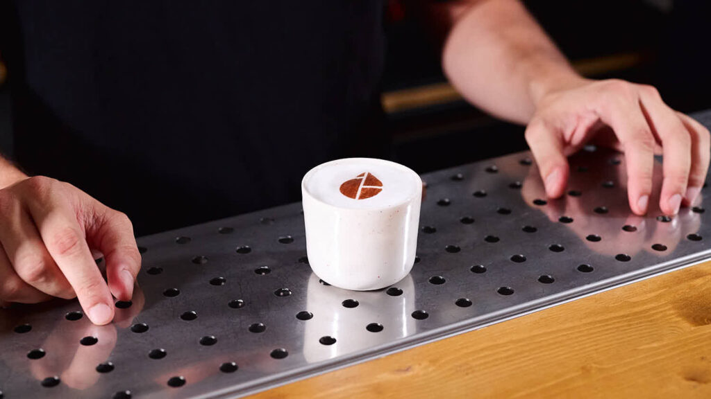 Su un banco bar un cocktail in stile sour (con schiumetta in suoerficie) servito all'interno di una mug in ceramica. Il cocktail è decorato con la stampa del logo Cocktail Engineering direttamente sulla superficie schiumosa