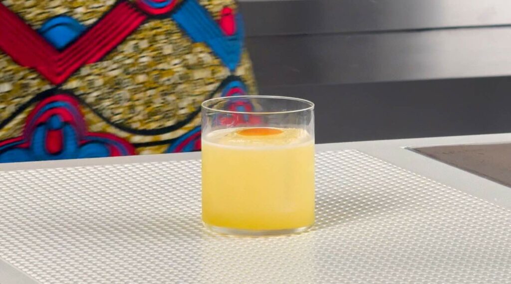 Su un banco bar il cocktail Rey Mysterio. Il cocktail si presenta di colore giallo, è servito in un bicchiere tumbler basso con un chunk di ghiaccio e decorato con una scorza di arancia