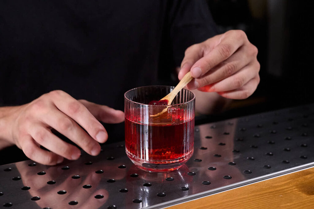 Su un banco bar un cocktail Negroni aromatizzato sfruttando la tecnica "sauce&jam washing" con confettura homemade alle arance rosse e ciliegie. Il drink è servito con un chunk di ghiaccio in un tumbler basso rigato ed è decorato con un cucchiaino in legno contenente la confettura