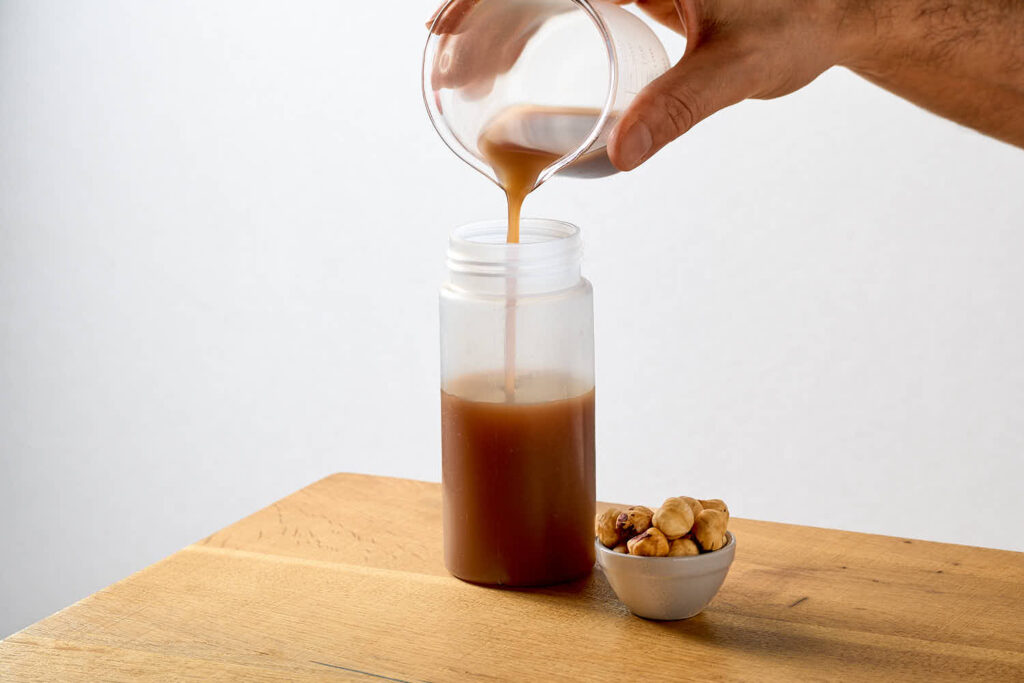 Su un tavolo in legno una bottiglietta squeezer dentro la quale viene versato (da un becker in vetro) lo sciroppo di nutella senza nutella: uno sciroppo per cocktail preparato home made home made con nocciole e cioccolato