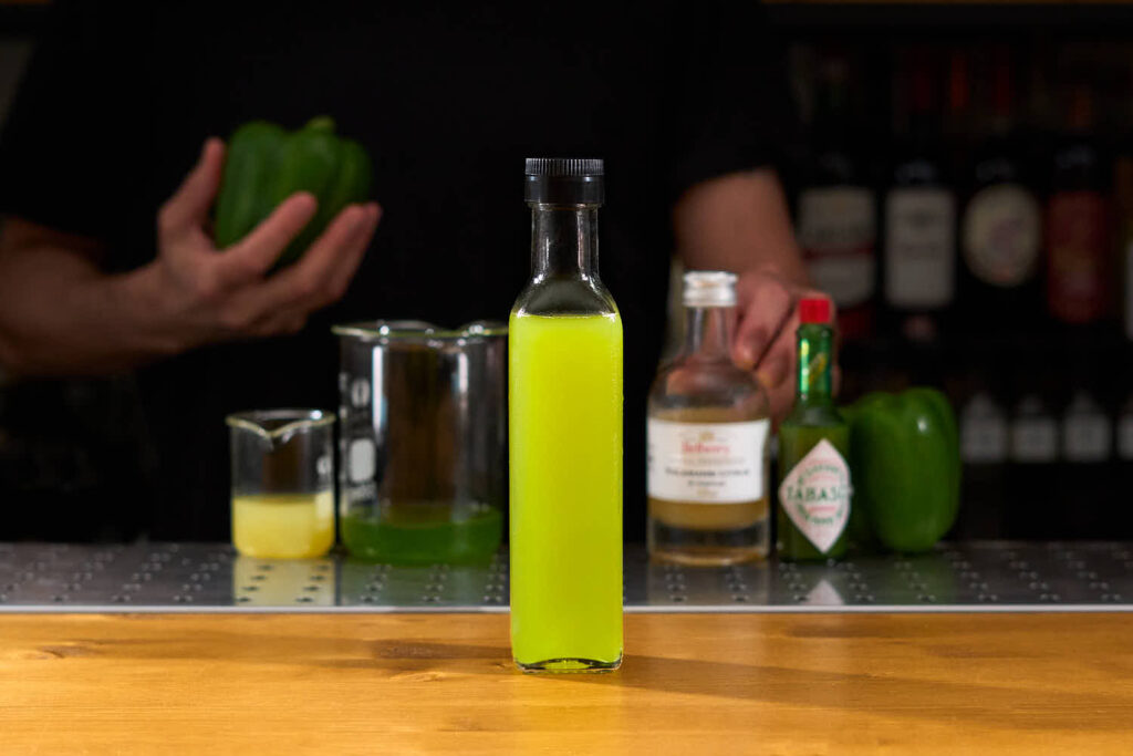 Su un banco bar una bottiglietta di vetro contenente una preparazione home made: shrub al peperone verde, tabasco verde, aceto al kalamansi, lime, e sciroppo di agave. Sullo sfondo gli ingredienti sfocati