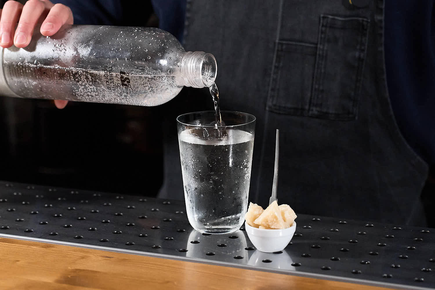 Su un banco bar un cocktail con soda alla pera servito in un bicchiere da long drink. Accanto al cocktail una piccola ciotola in ceramica bianca contenente dei pezzetti di Parmigiano reggiano. Si intravede la mano del barman che versa la soda nel bicchiere da un sifone 
